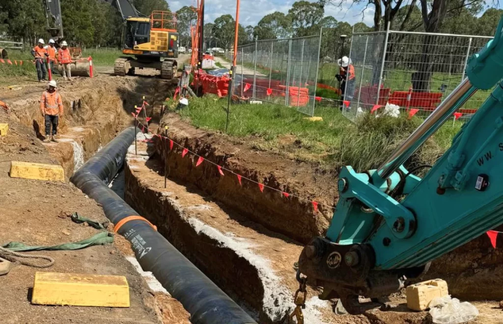 Construction workers install a large black pipe in a trench using heavy machinery, with safety barriers, fencing, and caution tape set up around the worksite.