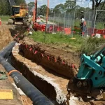 Construction workers install a large black pipe in a trench using heavy machinery, with safety barriers, fencing, and caution tape set up around the worksite.