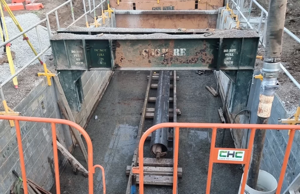 A construction site with a large steel beam above a trench, a pipe laid on wooden supports, and safety barriers surrounding the area.