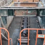 A construction site with a large steel beam above a trench, a pipe laid on wooden supports, and safety barriers surrounding the area.