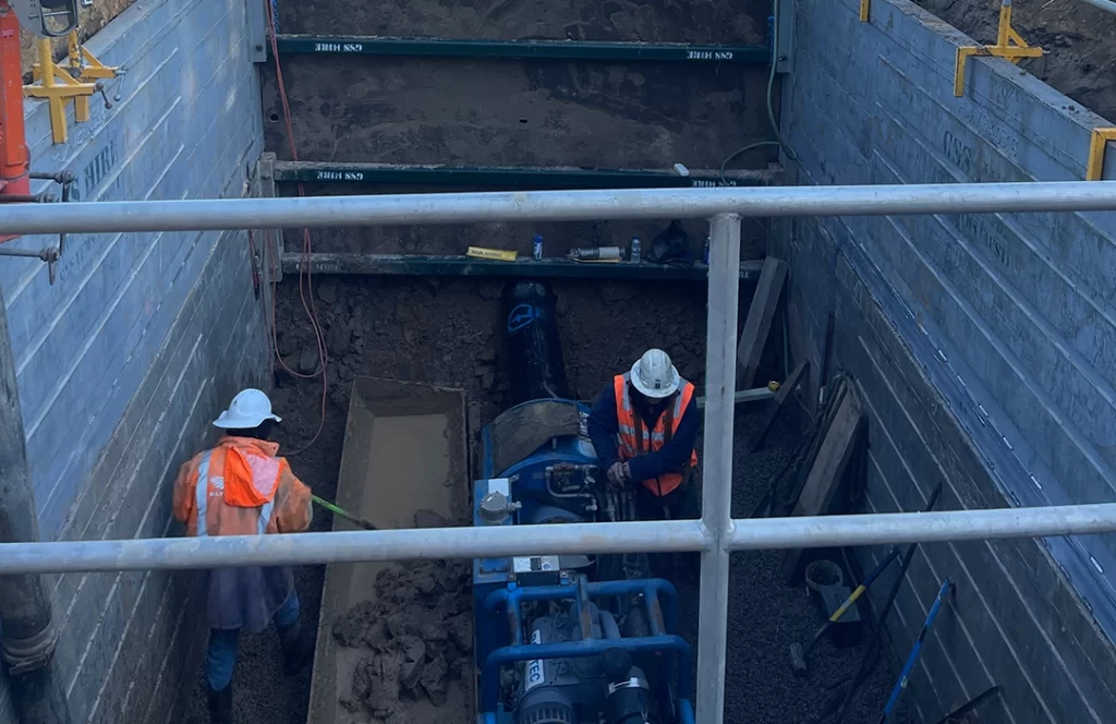 Two construction workers in safety gear work inside a reinforced excavation site with pipes, gravel, and equipment visible around them.