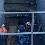 Two construction workers in safety gear work inside a reinforced excavation site with pipes, gravel, and equipment visible around them.