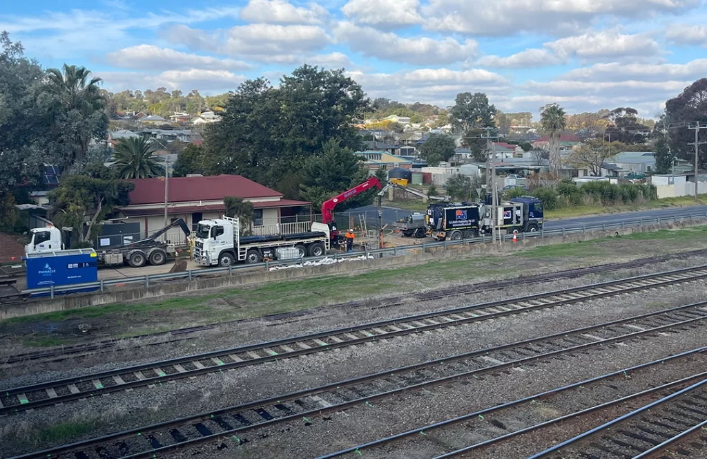 Two trucks and workers are positioned near a roadside construction site beside a set of railway tracks, with houses and trees in the background under a partly cloudy sky.