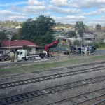 Two trucks and workers are positioned near a roadside construction site beside a set of railway tracks, with houses and trees in the background under a partly cloudy sky.
