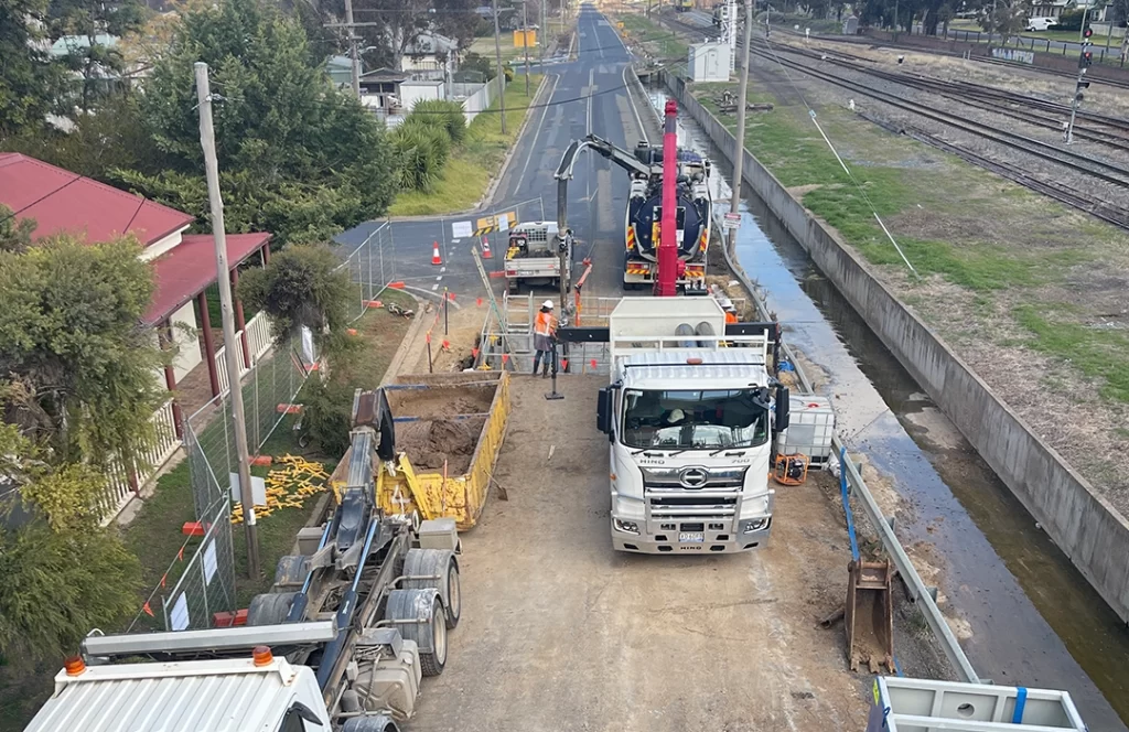 A construction site beside a railway features trucks, barriers, workers in safety gear, and construction equipment along a partially closed road.