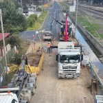 A construction site beside a railway features trucks, barriers, workers in safety gear, and construction equipment along a partially closed road.