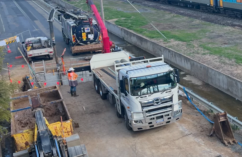 Construction workers and vehicles, including a white truck and excavator, are present at a work site near a railway track with safety barriers and equipment.