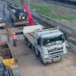 Construction workers and vehicles, including a white truck and excavator, are present at a work site near a railway track with safety barriers and equipment.