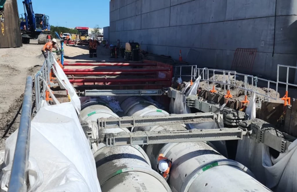 Construction workers install large concrete pipes in a trench beside a concrete wall, with machinery and safety barriers present at the site.
