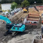 Excavators and workers operate on a construction site with large pipes, containers, and piles of sand and gravel under an overcast sky.