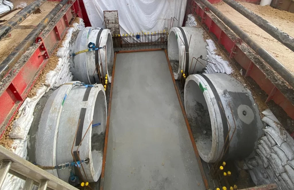 Four large concrete pipe sections are positioned horizontally within a rectangular excavation site lined with sandbags and metal supports.