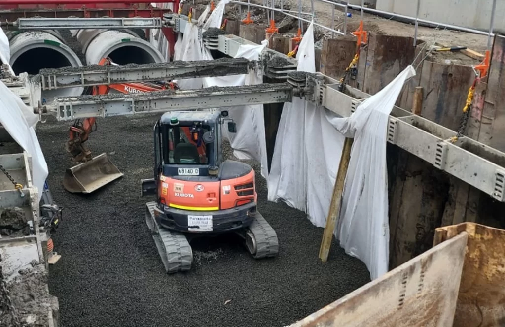 A small excavator operates in a trench lined with white sheets and gravel, with construction equipment and large concrete pipes visible in the background.