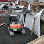 A small excavator operates in a trench lined with white sheets and gravel, with construction equipment and large concrete pipes visible in the background.