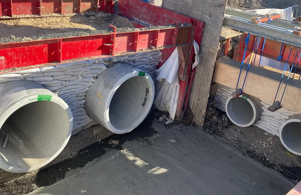 Three large concrete drainage pipes are installed in a construction site wall, surrounded by sandbags and forming part of a foundation structure.