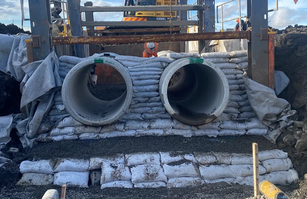Two large concrete pipes are installed side by side, supported by stacked sandbags, with construction equipment and a worker visible in the background.