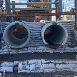 Two large concrete pipes are installed side by side, supported by stacked sandbags, with construction equipment and a worker visible in the background.