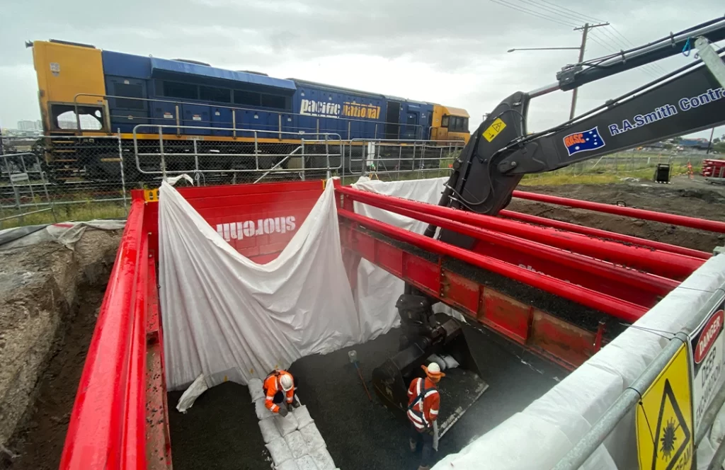 Two construction workers work inside a shored trench while an excavator operates above ground; a freight train passes by in the background behind a safety fence.