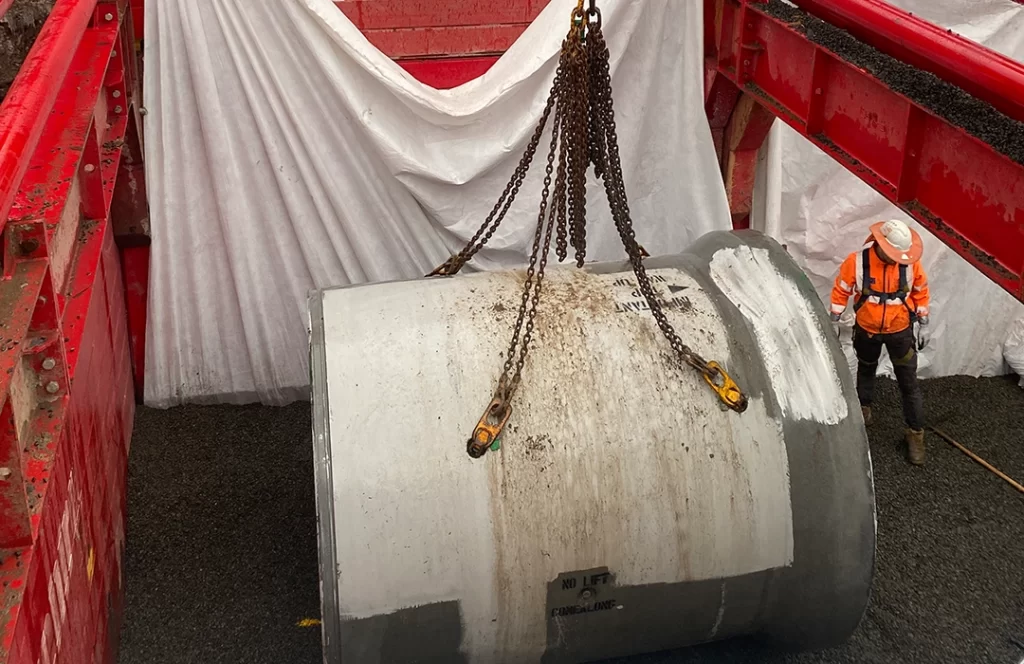 A large concrete pipe is lifted by chains from a crane, while a worker in high-visibility gear and a hard hat stands nearby on gravel inside a construction area.