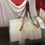 A large concrete pipe is lifted by chains from a crane, while a worker in high-visibility gear and a hard hat stands nearby on gravel inside a construction area.