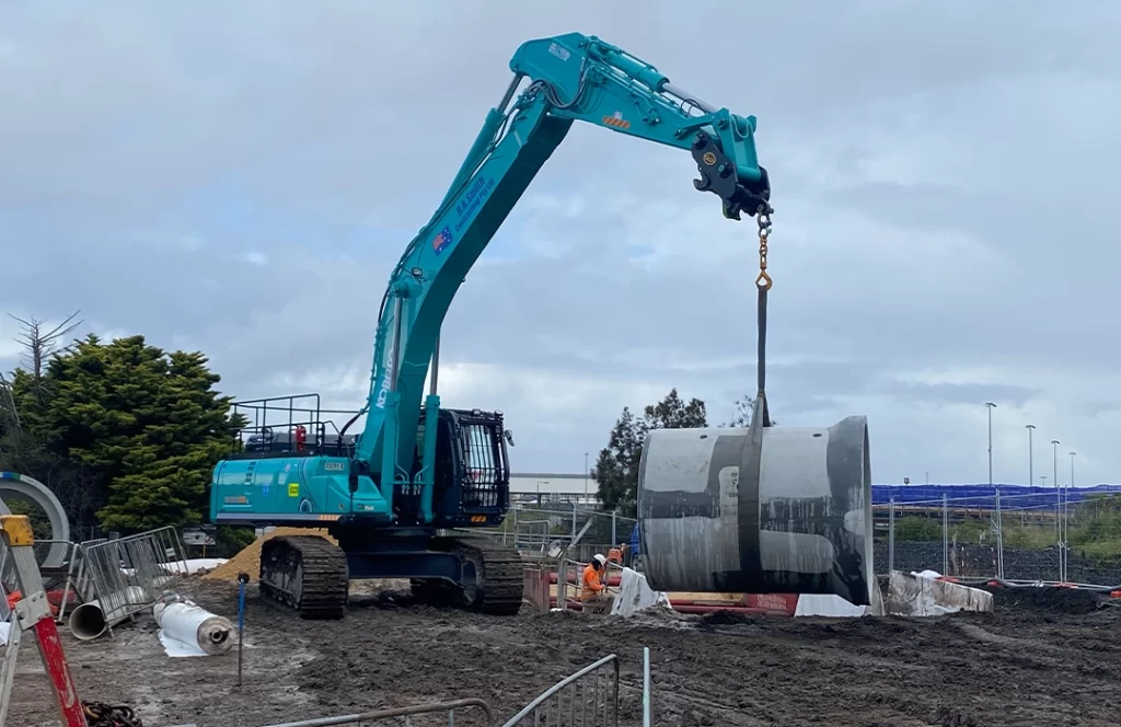 A teal excavator lifts a large concrete pipe at a muddy construction site, with workers and fencing visible in the background under a cloudy sky.
