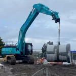 A teal excavator lifts a large concrete pipe at a muddy construction site, with workers and fencing visible in the background under a cloudy sky.
