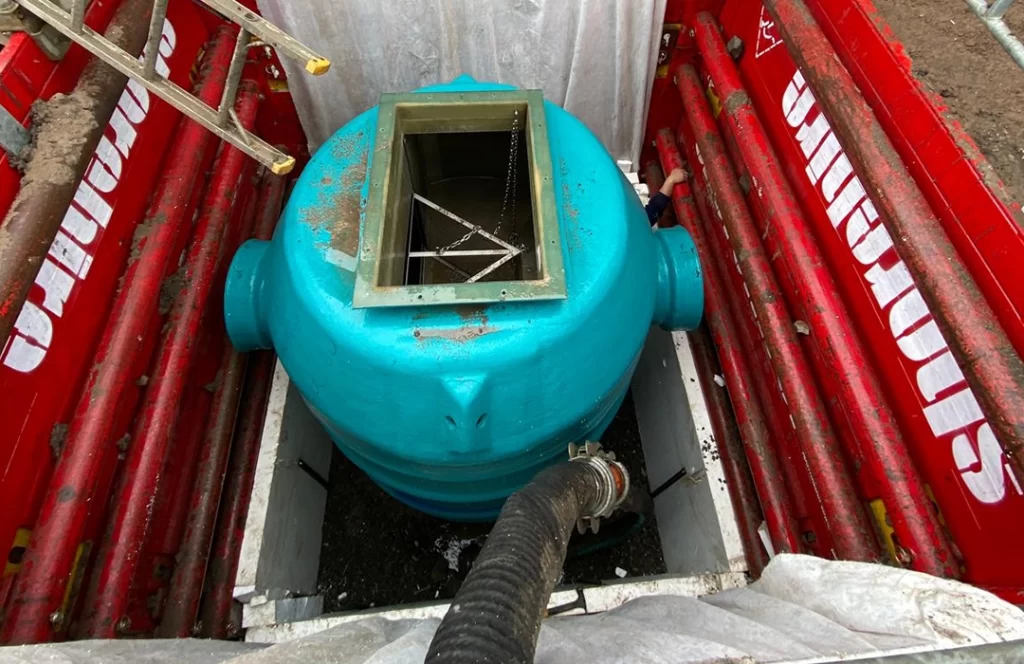 A blue cylindrical tank with a square open top is positioned in a red metal enclosure, with a black hose connected to its side.
