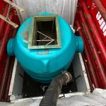 A blue cylindrical tank with a square open top is positioned in a red metal enclosure, with a black hose connected to its side.