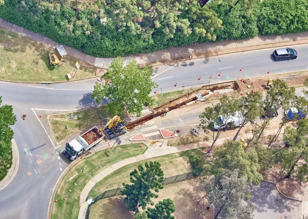 Aerial view of a road construction site with heavy machinery, trucks, and barriers, adjacent to a curved road and green trees.