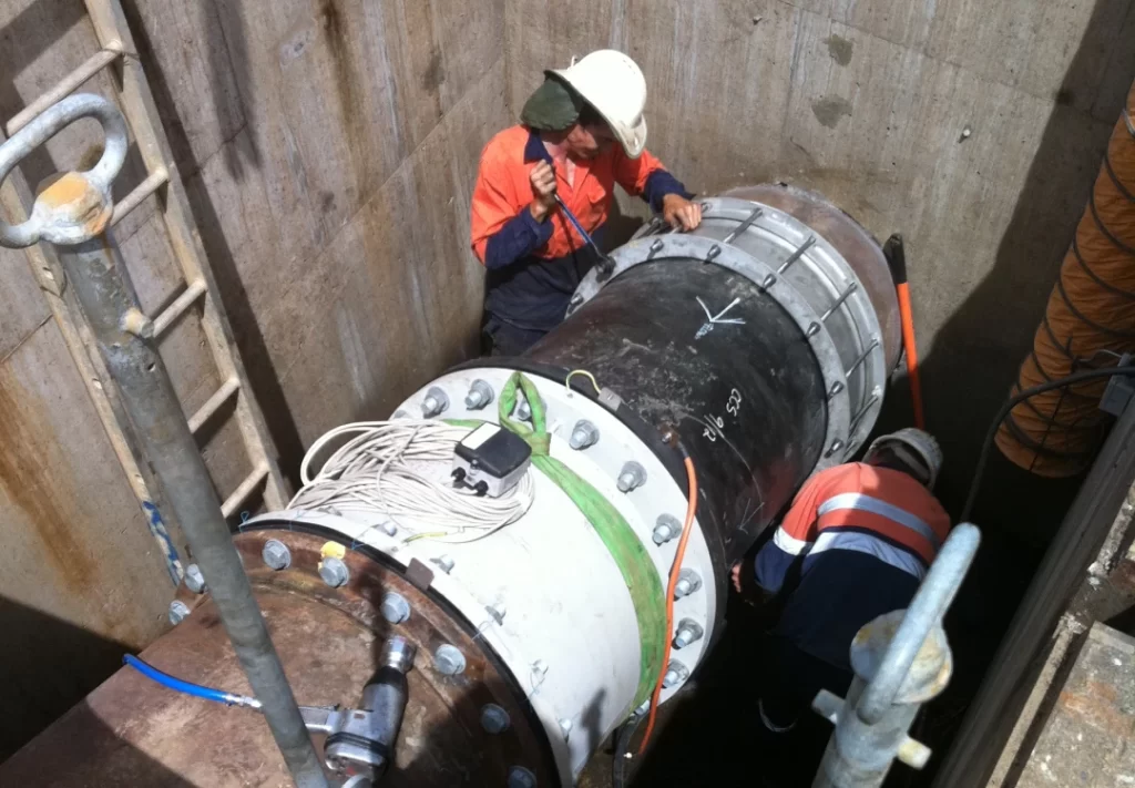 Two workers in safety gear inspect and work on a large industrial pipe in a concrete enclosure, with cables and equipment nearby.