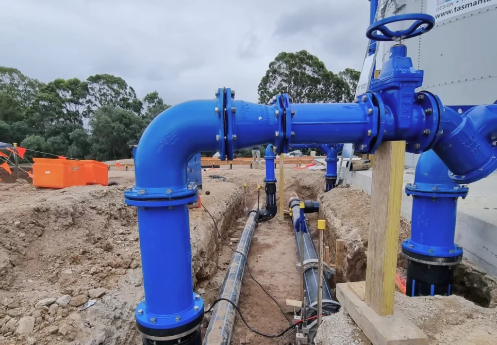 Large blue industrial water pipes are installed above and below ground at a construction site, with dirt, safety barriers, and trees in the background.