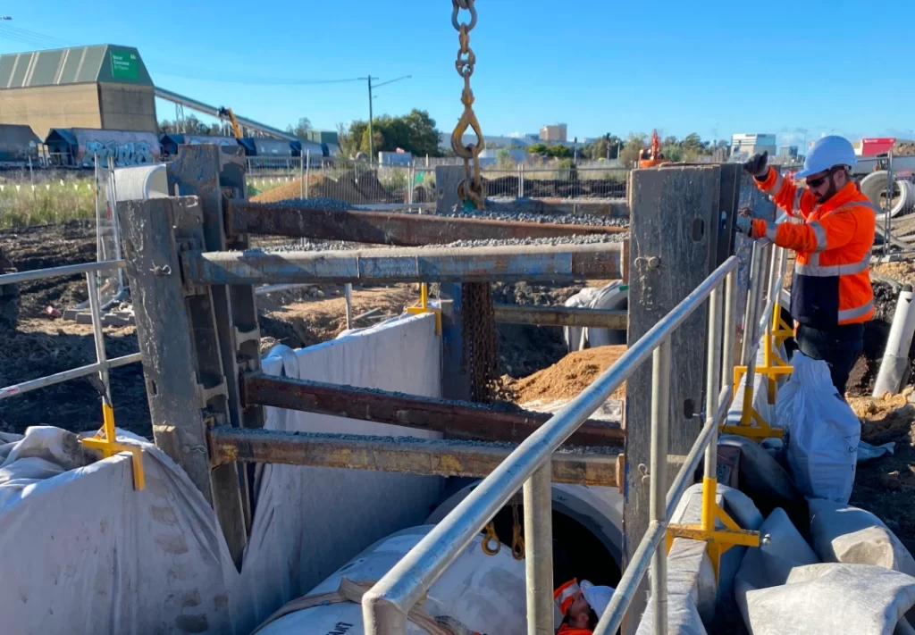 A construction worker in safety gear operates next to a large metal support structure over a trench with pipes and construction materials visible.