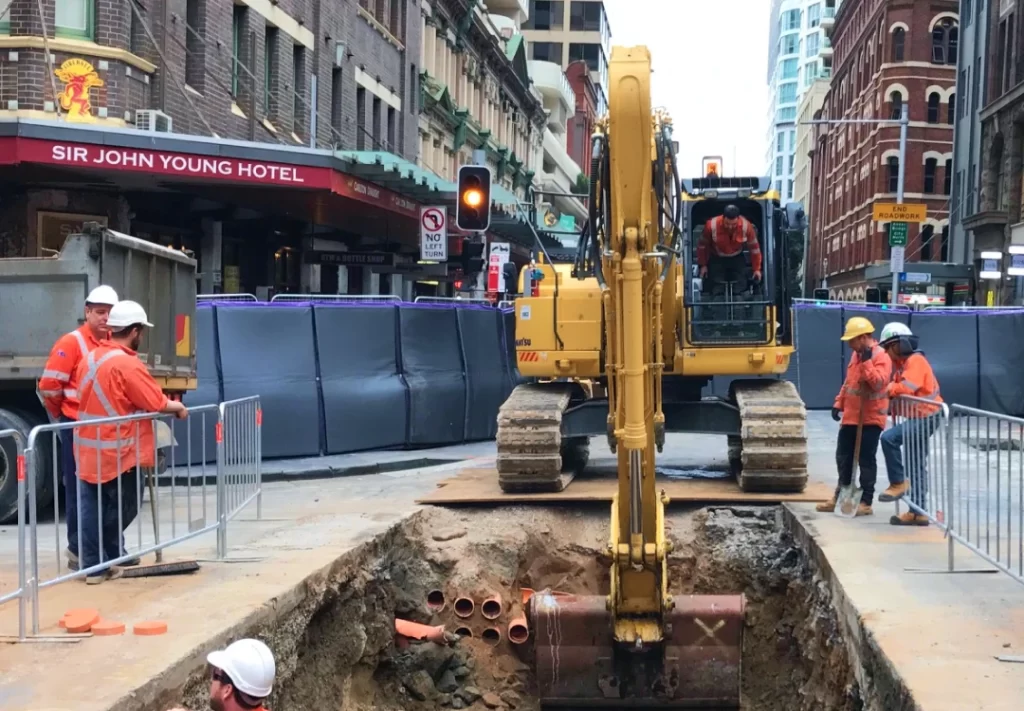 Construction workers in safety gear stand near an excavator digging a trench with exposed pipes on a city street, surrounded by barriers and buildings.