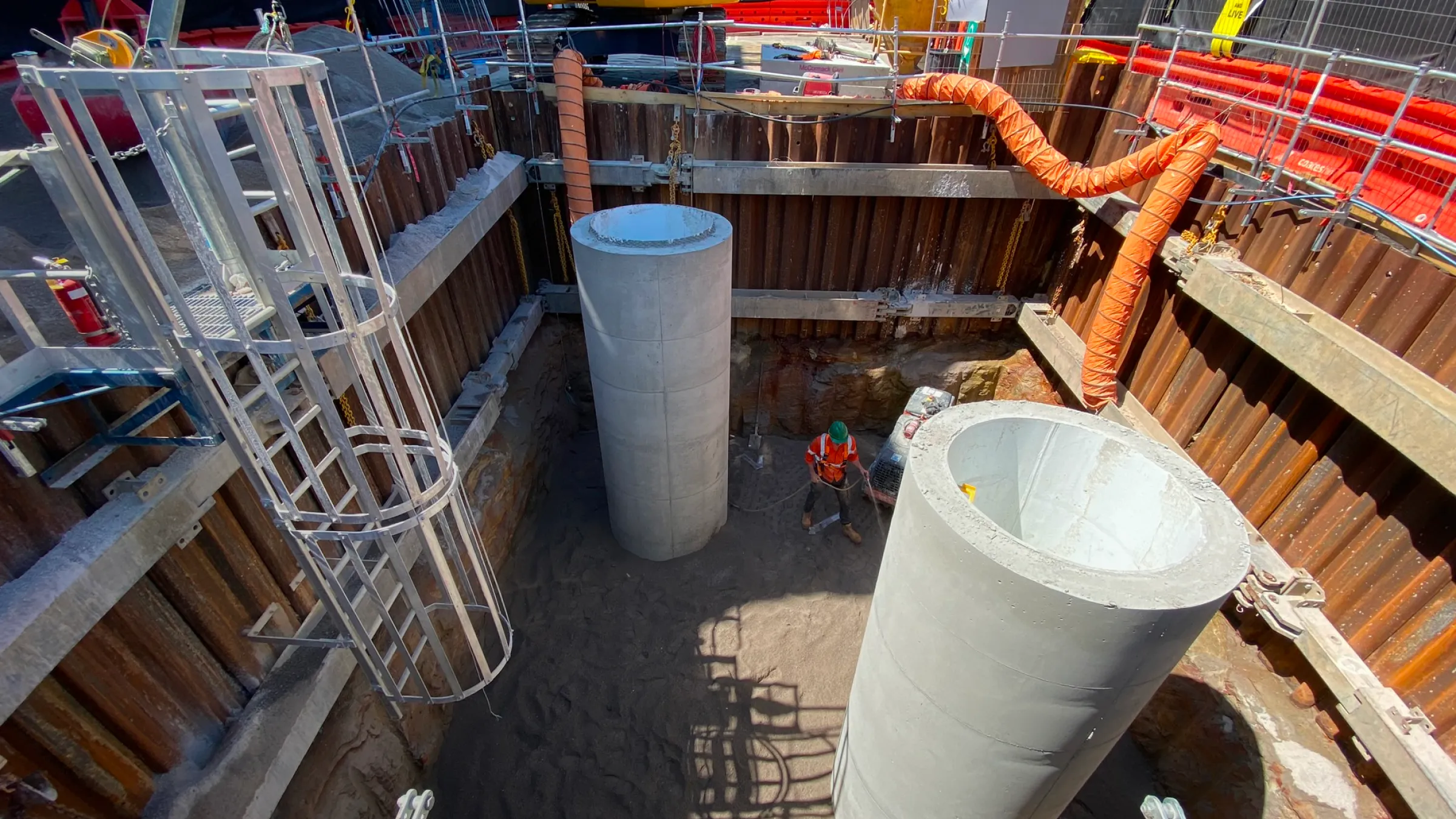 Construction site with two large concrete cylinders, metal ladder, orange ventilation ducts, and a worker in a helmet inside an excavated area.