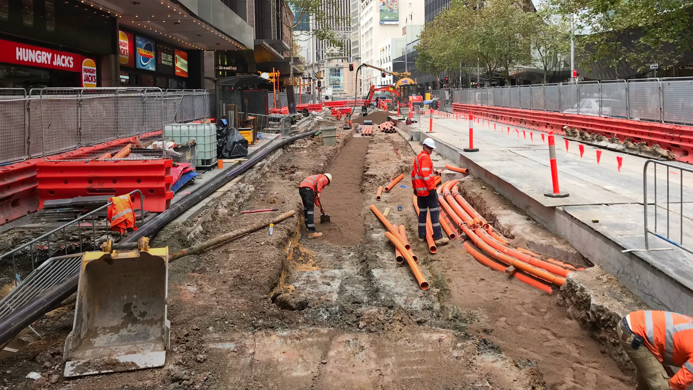 Construction workers in high-visibility gear install orange utility pipes in a fenced-off urban street undergoing major roadwork and excavation.