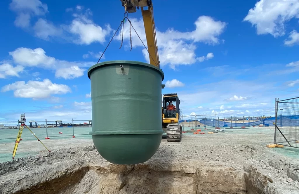 A construction vehicle lifts a large green cylindrical tank over a dirt excavation site under a partly cloudy sky.