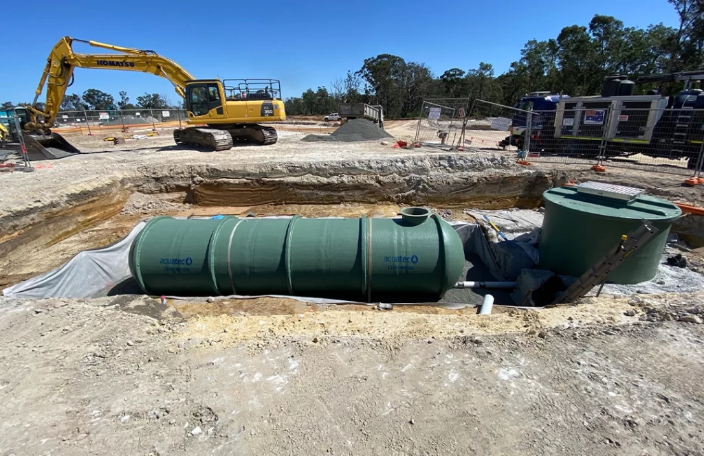 A large green underground water tank is being installed at a construction site, with an excavator and construction materials visible in the background.