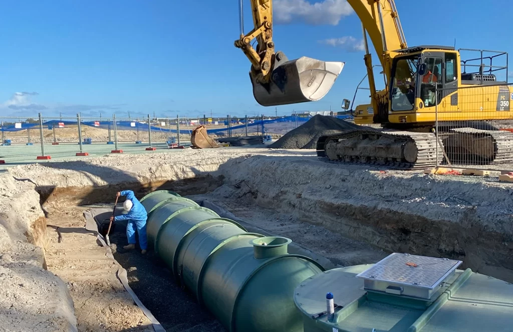 Construction worker in blue coveralls installs large green storage tanks in a trench as a yellow excavator operates nearby on a construction site.