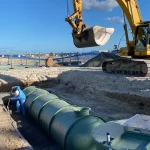 Construction worker in blue coveralls installs large green storage tanks in a trench as a yellow excavator operates nearby on a construction site.