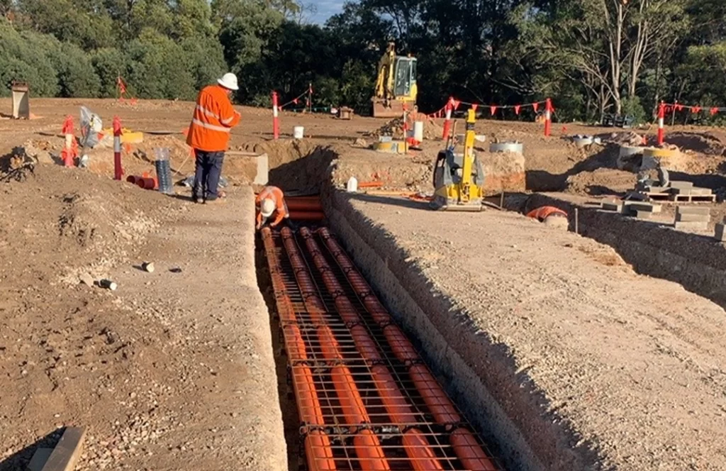 Construction workers in high-visibility clothing install orange utility pipes in a trench, surrounded by dirt and construction equipment.