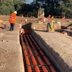 Construction workers in high-visibility clothing install orange utility pipes in a trench, surrounded by dirt and construction equipment.