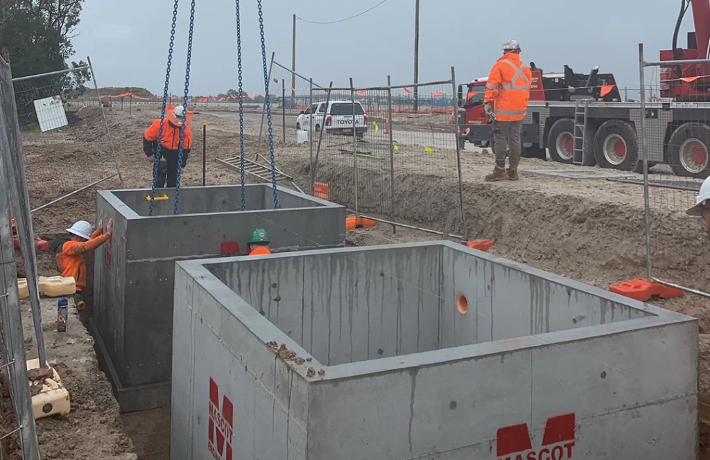 Construction workers in high-visibility clothing install large concrete utility boxes in a trench at a worksite using a crane. Safety barriers and vehicles are visible in the background.