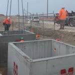 Construction workers in high-visibility clothing install large concrete utility boxes in a trench at a worksite using a crane. Safety barriers and vehicles are visible in the background.