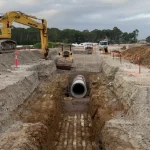 Excavators and construction vehicles work at a construction site with a large concrete pipe being installed in a deep trench.