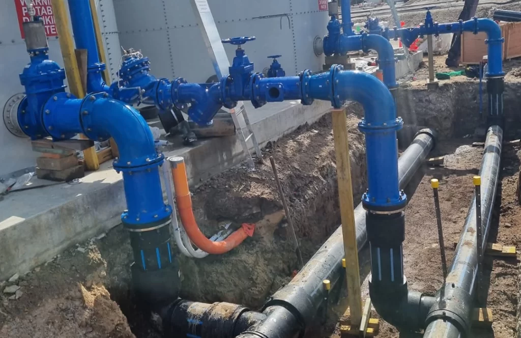 A series of blue industrial pipes and valves connected to a building, with additional black and orange pipes running underground in a trench.