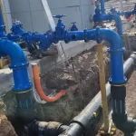 A series of blue industrial pipes and valves connected to a building, with additional black and orange pipes running underground in a trench.