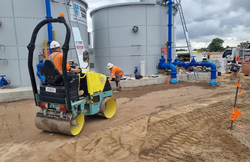 Two construction workers in safety gear work near large metal tanks; one operates a small road roller on a dirt surface, while the other works near blue pipes and site equipment.