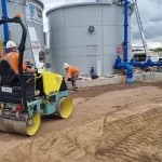 Two construction workers in safety gear work near large metal tanks; one operates a small road roller on a dirt surface, while the other works near blue pipes and site equipment.