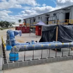 Industrial site with large pipes wrapped in blue plastic, concrete barriers, safety railings, and a building with yellow accents in the background under a partly cloudy sky.