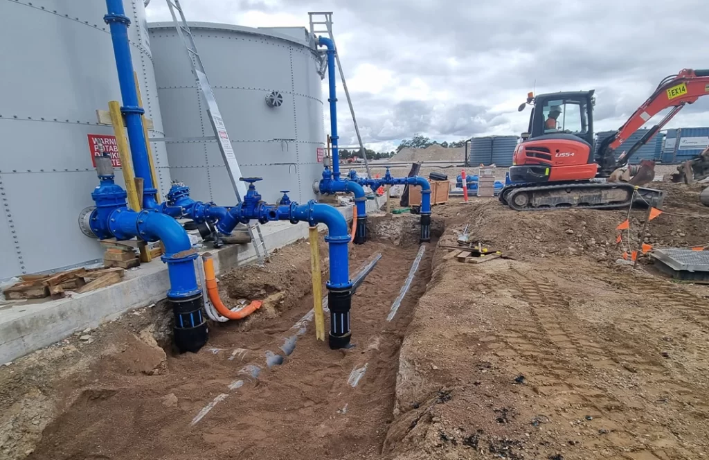 A construction site with blue industrial pipes connected to a large storage tank, an orange excavator, and exposed ground.