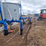 A construction site with blue industrial pipes connected to a large storage tank, an orange excavator, and exposed ground.